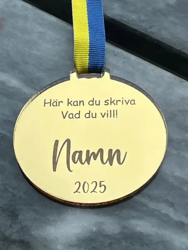 Medalj | Skriv vad du vill av Express-presenter: En guldmedalj med blågult sverigeband, svensk gravyr "Här kan du skriva Vad du vill! Namn 2025", tillverkad av spegelplast, visas på en grå yta.