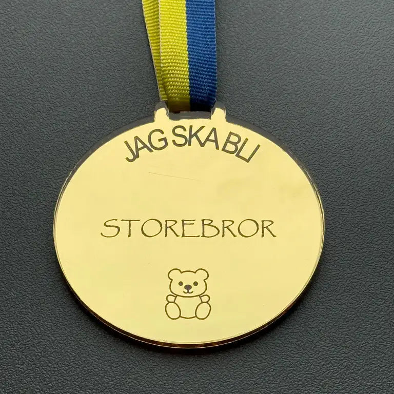 Medalj | Jag ska bli storebror från Express-presenter är en guldfärgad spegelsplastmedalj med blågult band, med texten "JAG SKA BLI STOREBROR" ovanför en nallebjörn, visad på en mörk, strukturerad yta.