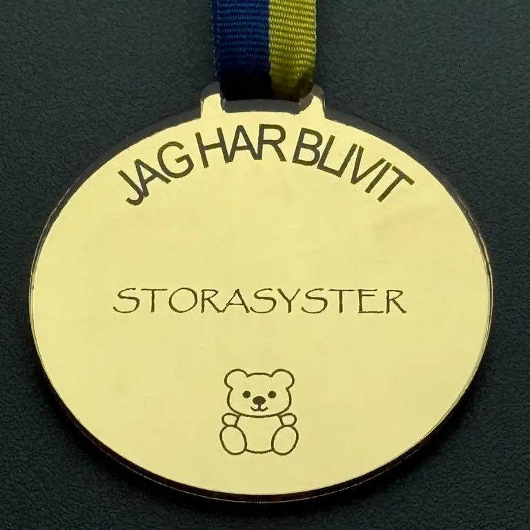 Medalj | Jag har blivit storasyster" från Express-presenter har en guldmedalj med ett blått och gult band och en söt nallebjörn - perfekt för att fira nya storasystrar.