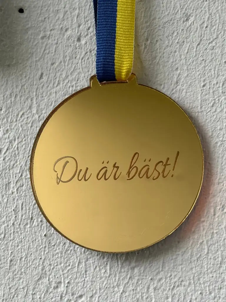 Medalj | Du är bäst från Express-presenter är en guldfärgad spegelplastmedalj med "Du är bäst!"-gravyr, blått och gult band, lasergraverad och svensktillverkad. På bilden hängande på en vit strukturerad vägg.