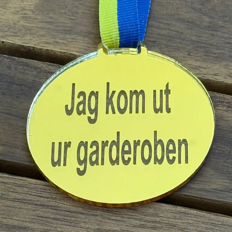 Express-presenter Medalj - Jag kom ut ur garderoben är en gul oval medalj i Spegelplast guld, 70 x 3 mm, med blågult band och svensk text, vilande på en träyta.