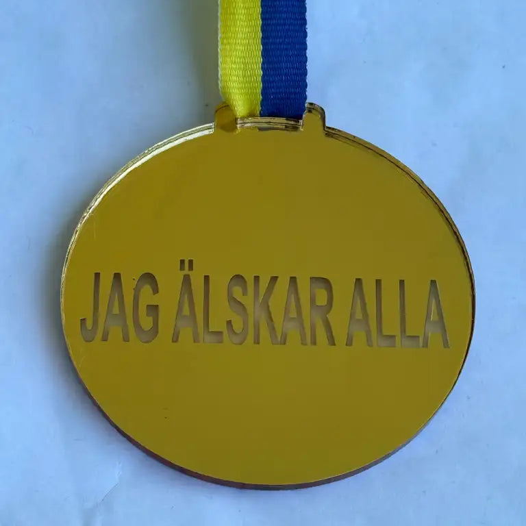 Express-presenter Medalj - Jag älskar alla är en guldmedalj i spegelplast med "JAG ÄLSKAR ALLA" ingraverat, fäst vid ett blågult band (70 x 3 mm), avbildad liggande på en vit yta.