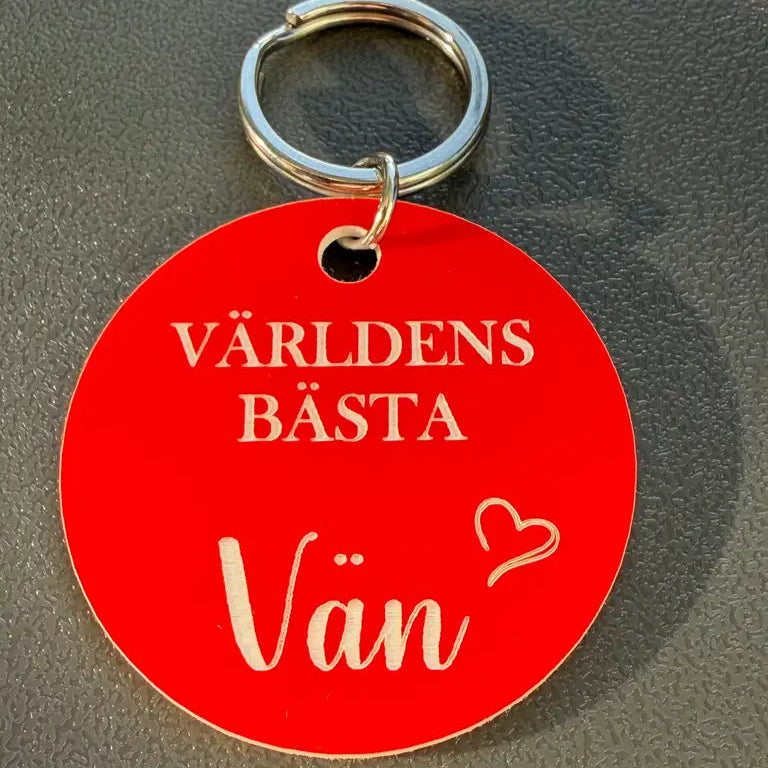 Nyckelring | Världens bästa vän (Röd) från Express-presenter - en rund, röd graverad nyckelring med svensk text och litet hjärta, perfekt som present till bästa vän. Ligger på en grå yta.