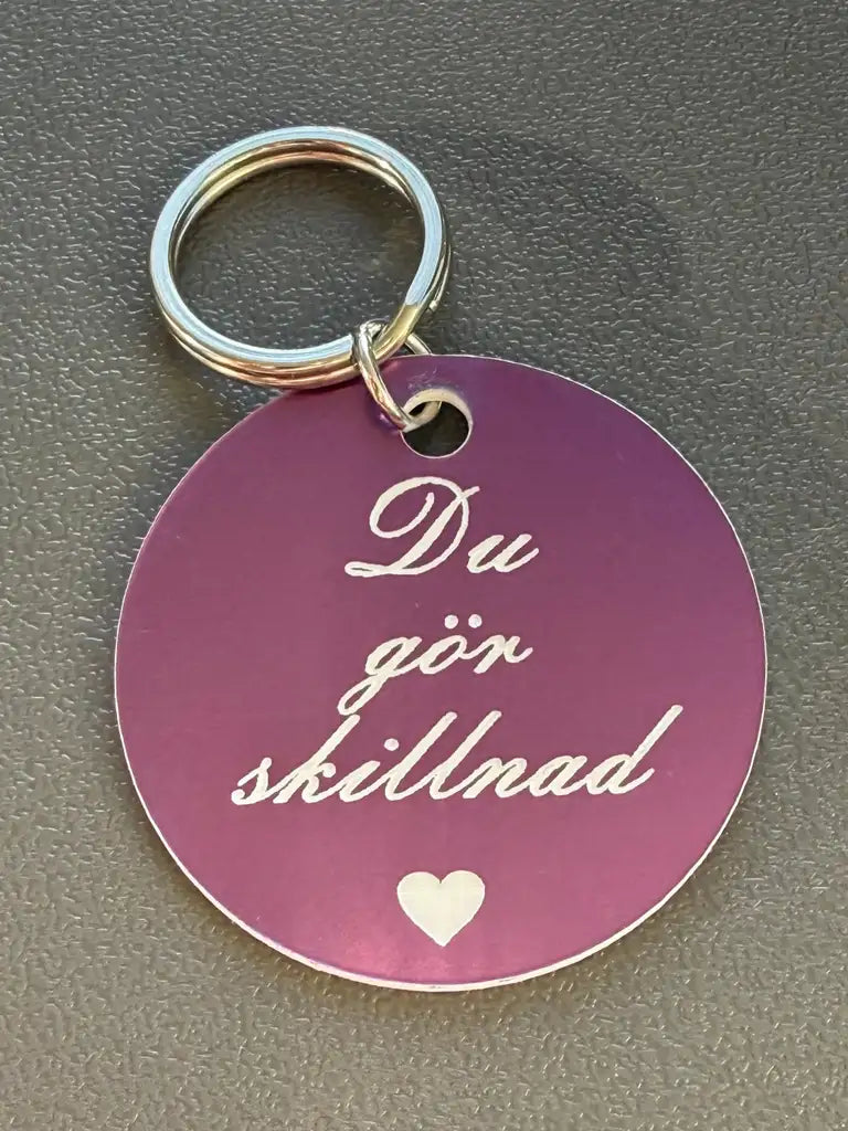 Nyckelring | Du gör skillnad från Express-presenter: Rund, mörklila nyckelring (40 mm) med silvertext "Du gör skillnad" och ett vitt hjärta under, fotograferad på en texturerad grå yta.