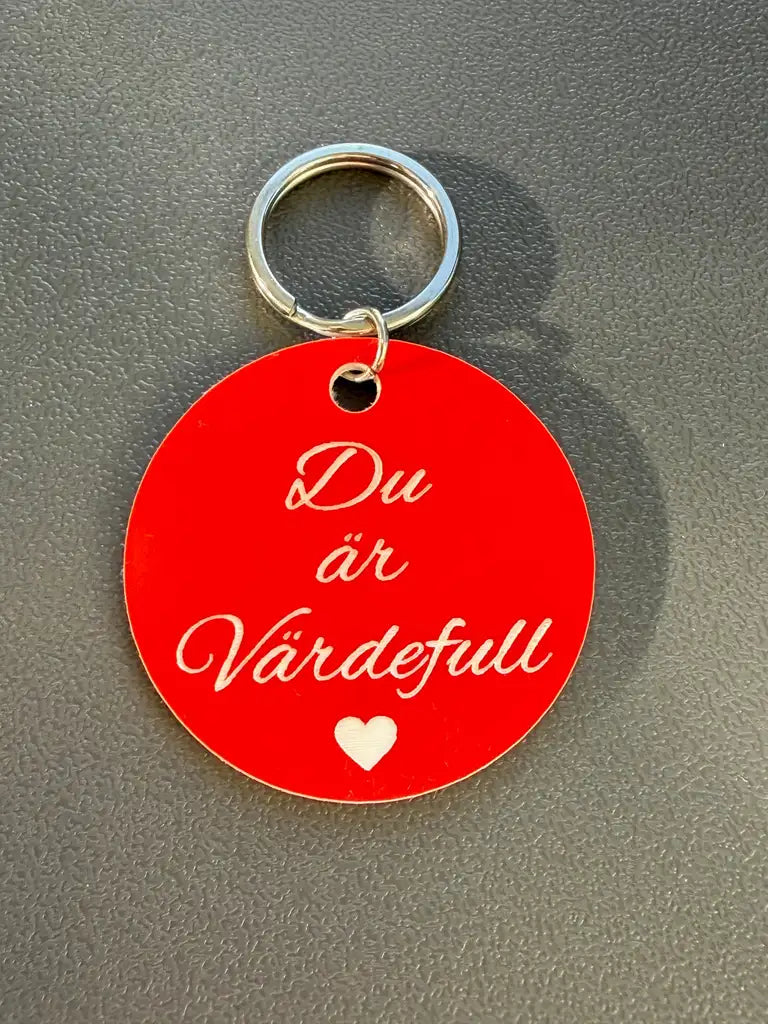 Nyckelring "Du är värdefull (röd)" från Express-presenter: 40mm bred röd nyckelring med svensk vit text "Du är Värdefull" och ett litet vitt hjärta undertill, placerad på en grå yta.