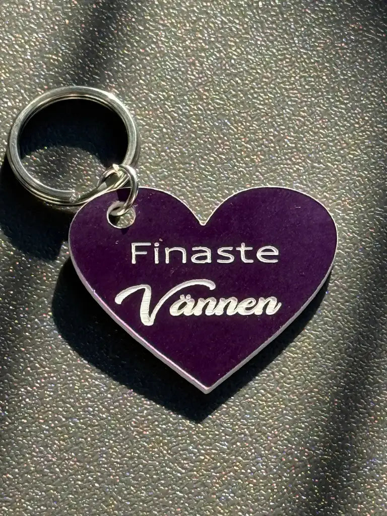 The "Nyckelring | Finaste vännen (lila)" from Express-presenter är en hjärtformad, lila nyckelring med texten "Finaste Vännen" och ligger på en grå, texturerad yta.
