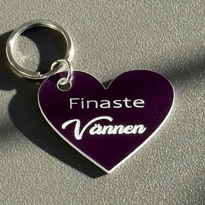 Nyckelring | Finaste vännen (lila) från Express-presenter, hjärtformad med texten "Finaste Vännen" graverad, ligger på en grå yta med mjuka skuggor i solljus.