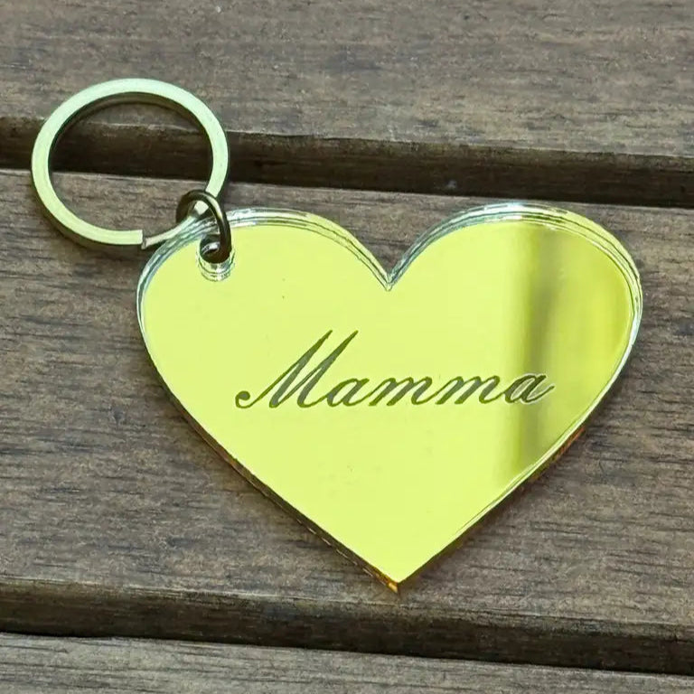 Nyckelring - Mamma by Express-presenter är en guldfärgad, hjärtformad nyckelring av 3mm spegelplast, graverad med "Mamma". Storlek: 4x3cm. Den vilar på en träyta.
