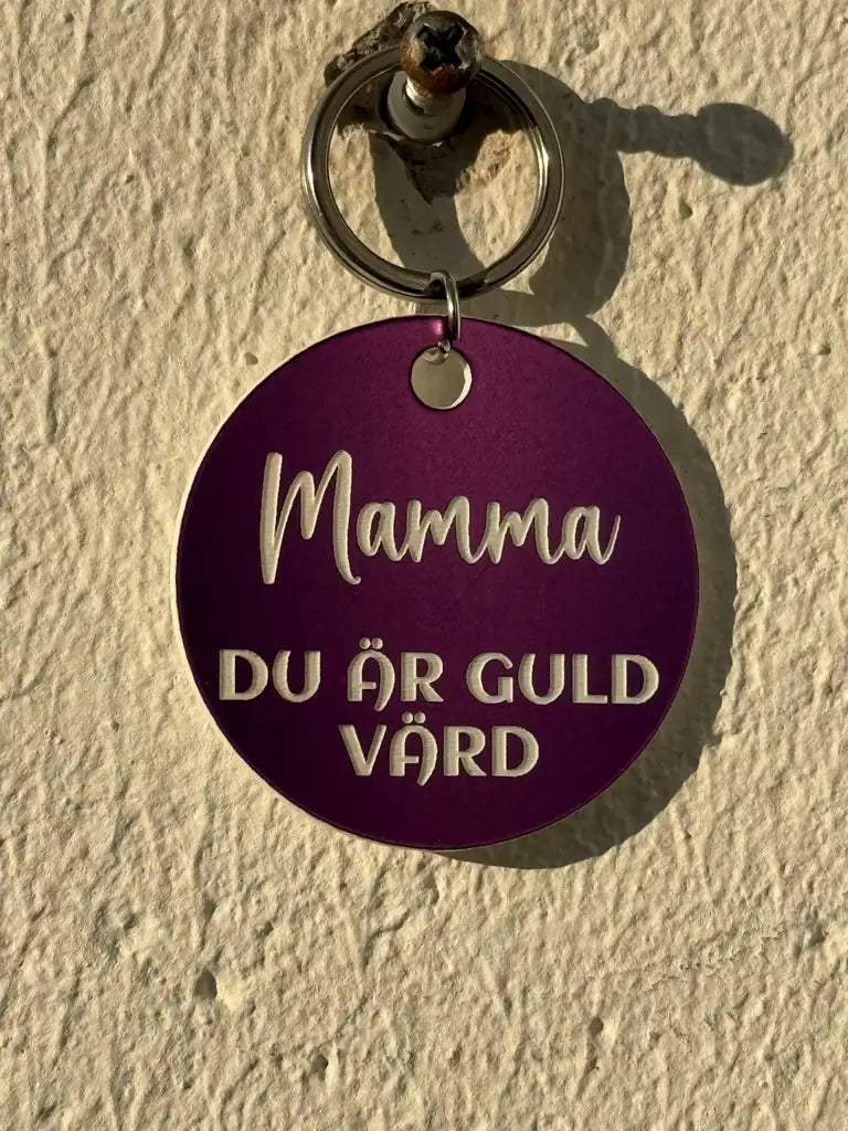 The Nyckelring | Mamma du är guld värd (Lila) från Express-presenter är en rund mörklila plastnyckelring med vit graverad text, upphängd mot en solbelyst, texturerad vägg.