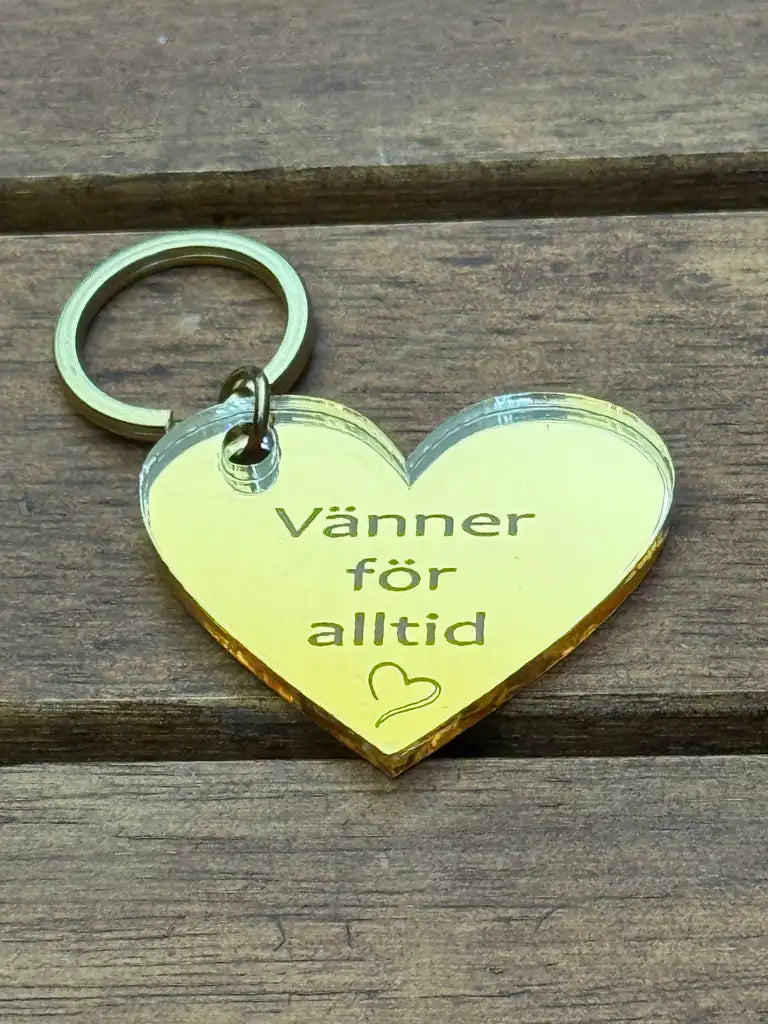 Nyckelring - Vänner för alltid från Express-presenter: en hjärtformad nyckelring i 3mm spegelplast (4x3 cm) med texten "Vänner för alltid" och ett litet hjärta, placerad på ett trägolv.