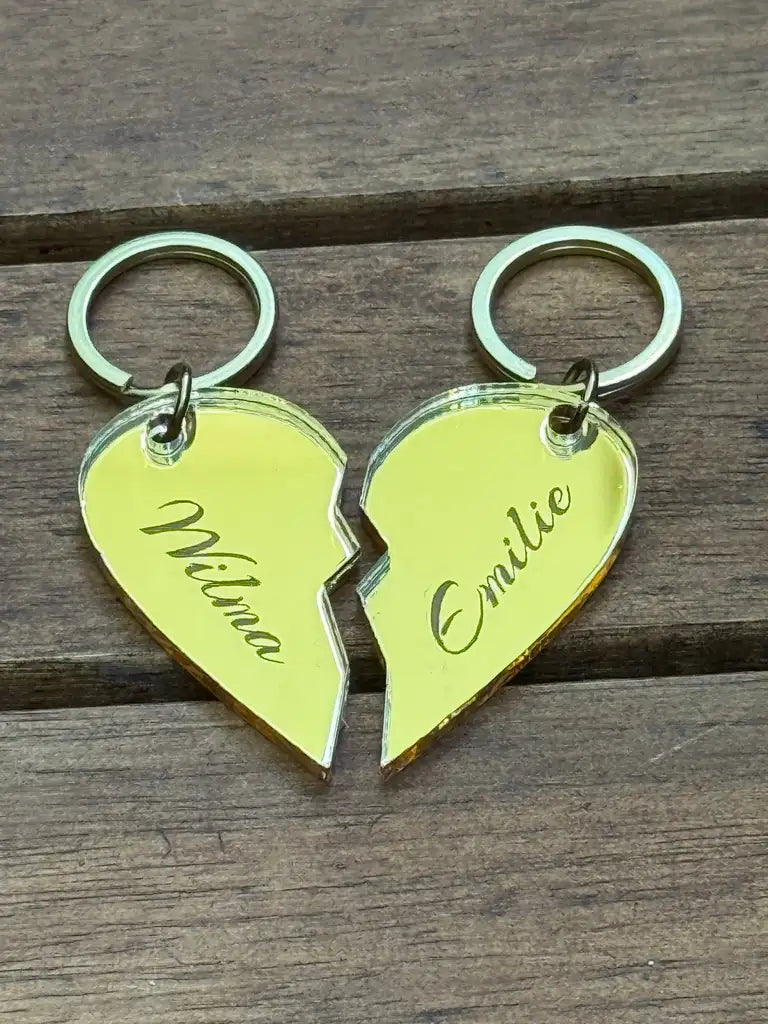 Två guldfärgade Nyckelring - Halvt hjärta-nyckelringar från Express-presenter, vardera 4x3cm, skapar ett namn i hjärta när de sätts ihop; "Milina" visas till vänster och "Emilie" till höger, visas på en träyta.