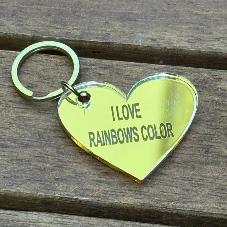 En guldfärgad, hjärtformad "Nyckelring - I love rainbows color" från Express-presenter, tillverkad av 3 mm spegelplast (4x3 cm) med graveringen "I LOVE RAINBOWS COLOR", ligger på en träyta.