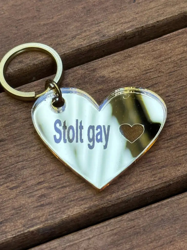 Express-presenter's "Nyckelring - Stolt gay" är en 4x3cm guldhjärta nyckelring i 3mm spegelplast, graverad med "Stolt gay" och ett litet hjärta. Den visas vilande på trä.