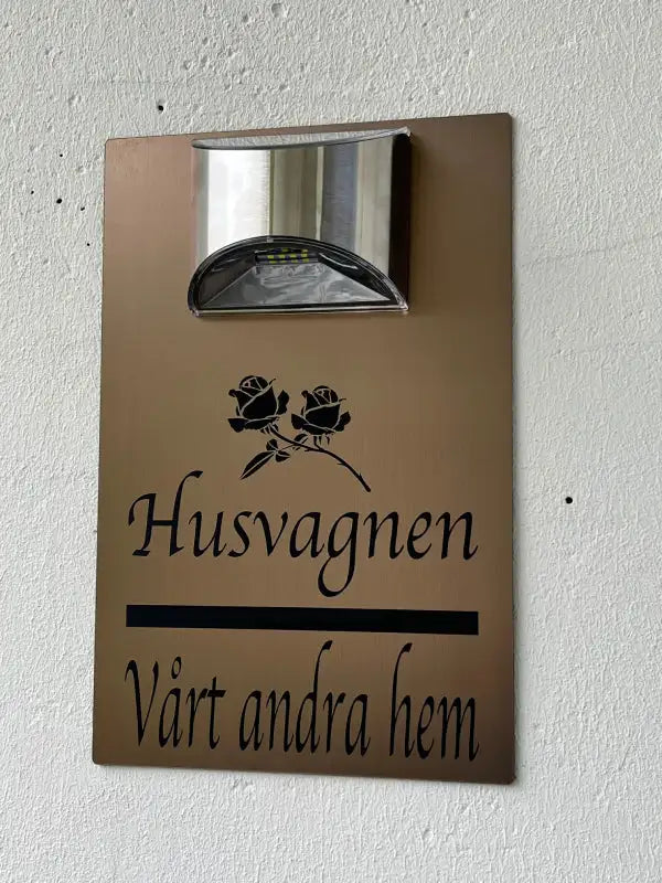 Expressens skylt Skylt till husvagnen visar en solcellslampa ovanför svarta blomstermotiv och den svenska texten "Husvagnen" och "Vårt andra hem", monterad på en vit vägg.
