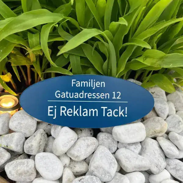 En blå oval Stilig adresskylt till Brevlådan by Express-presenter, tillverkad i akryl, pryder gröna växter och vita stenar. Påskriften är på svenska: Familjen Gatuadressen 12 Ej Reklam Tack! och ber artigt om att slippa annonser.