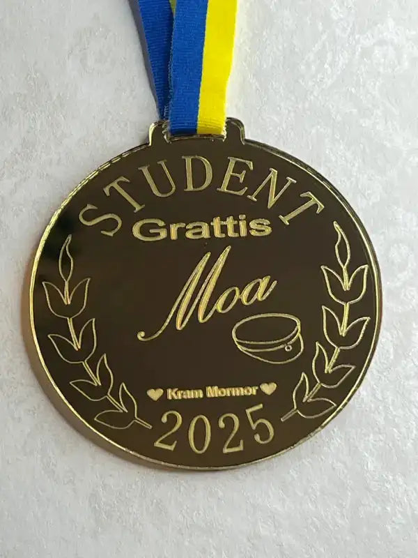 Express-presenter's Studentpaketet innehåller en studentmedalj i guld med blågult band. Texten lyder "STUDENT Grattis Moa Kram Mormor 2025" med motiv av lagerblad, hjärta och examensmössa - perfekt för studentpresenter.
