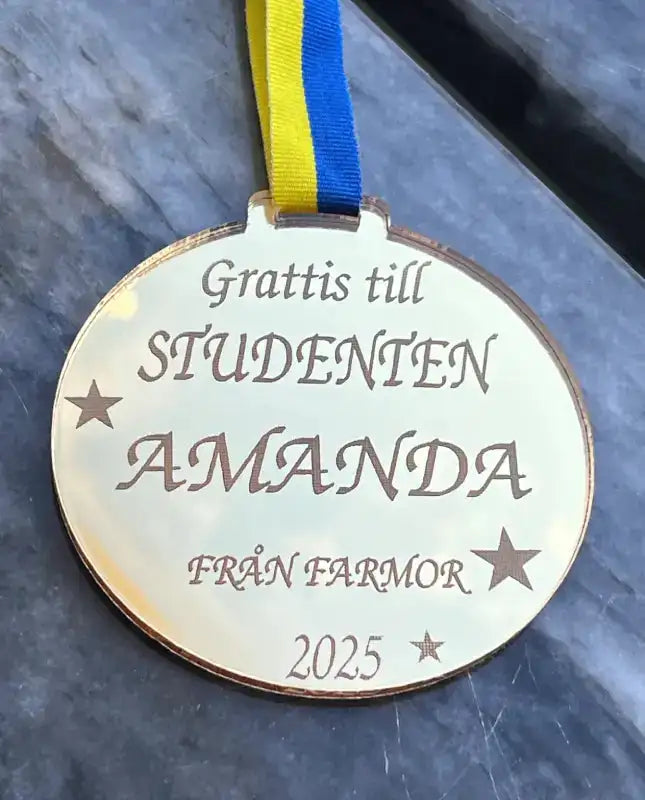 Express-presenter's Medalj | Studenten medalj med hälsning har blå-gult band och text: Grattis till STUDENTEN AMANDA FRÅN FARMOR 2025, på grå träyta-en perfekt personlig present.